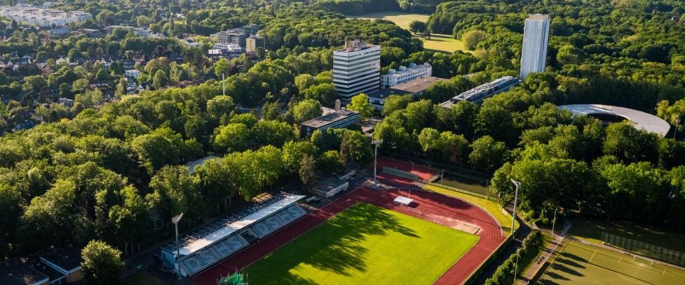 Eine Luftaufnahme des Campus der deutschen Sporthochschule Köln mit Stadion, Sportgelände, Instituts- und Verwaltungsgebäuden, umgeben von viel Baumbestand.